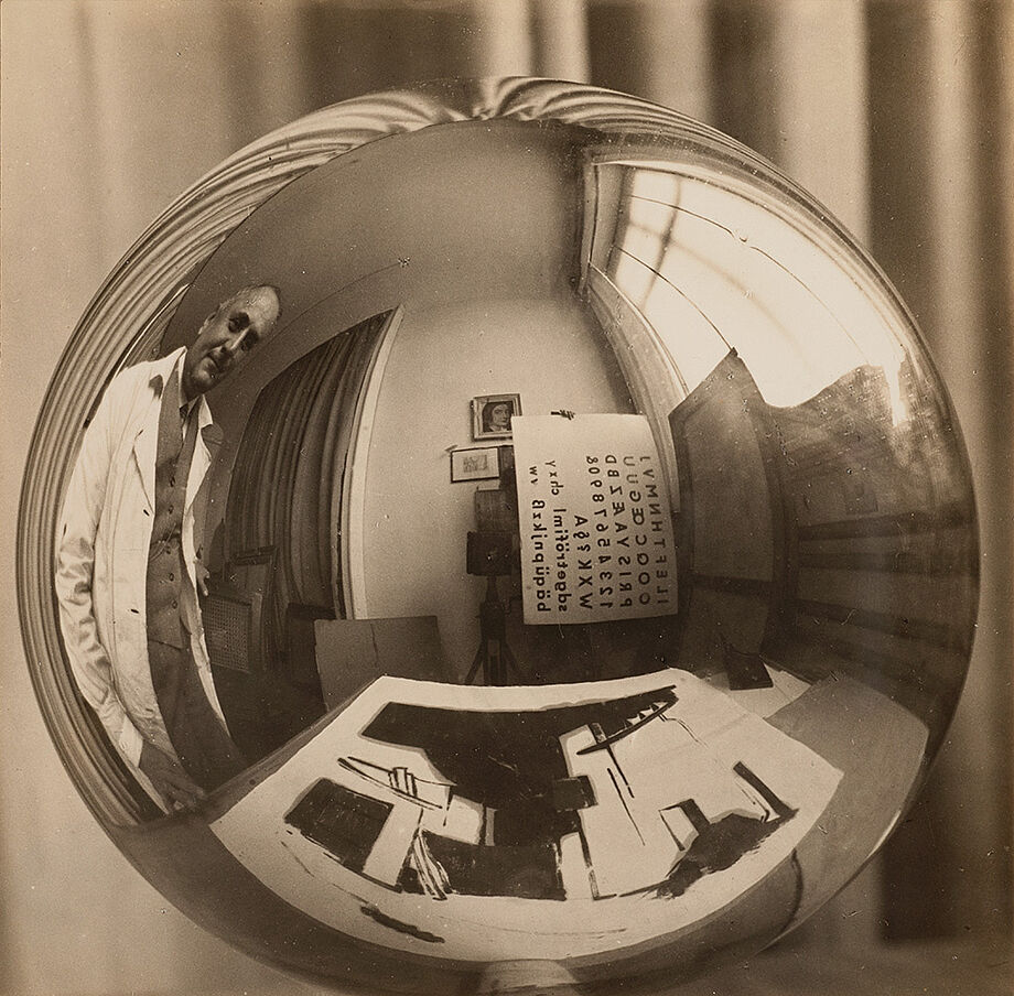 Das Bild zeigt Arno Drescher in seinem Atelier. Der Raum ist in einer Kugel gespiegelt und die Perspektive verzogen. Man sieht eine Zeichnung und eine Schriftart im Hintergrund.