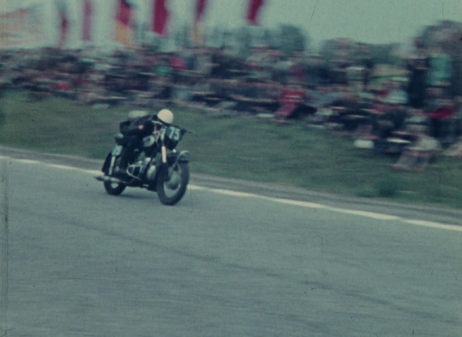 Ein Motorrad fährt über eine Rennstrecke. An der Seite stehen (verschwommen) jubelnde Zuschauer:innen.