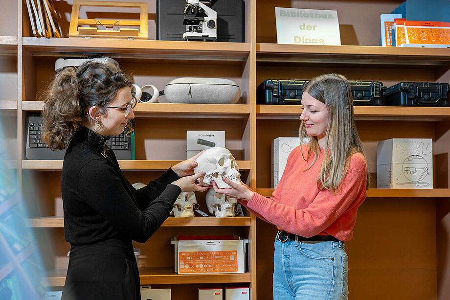 Zwei Personen mit anatomischem Modell eines menschlichen Schädels vor Regal mit weiteren Objekten aus der Bibliothek der Dinge in der SLUB